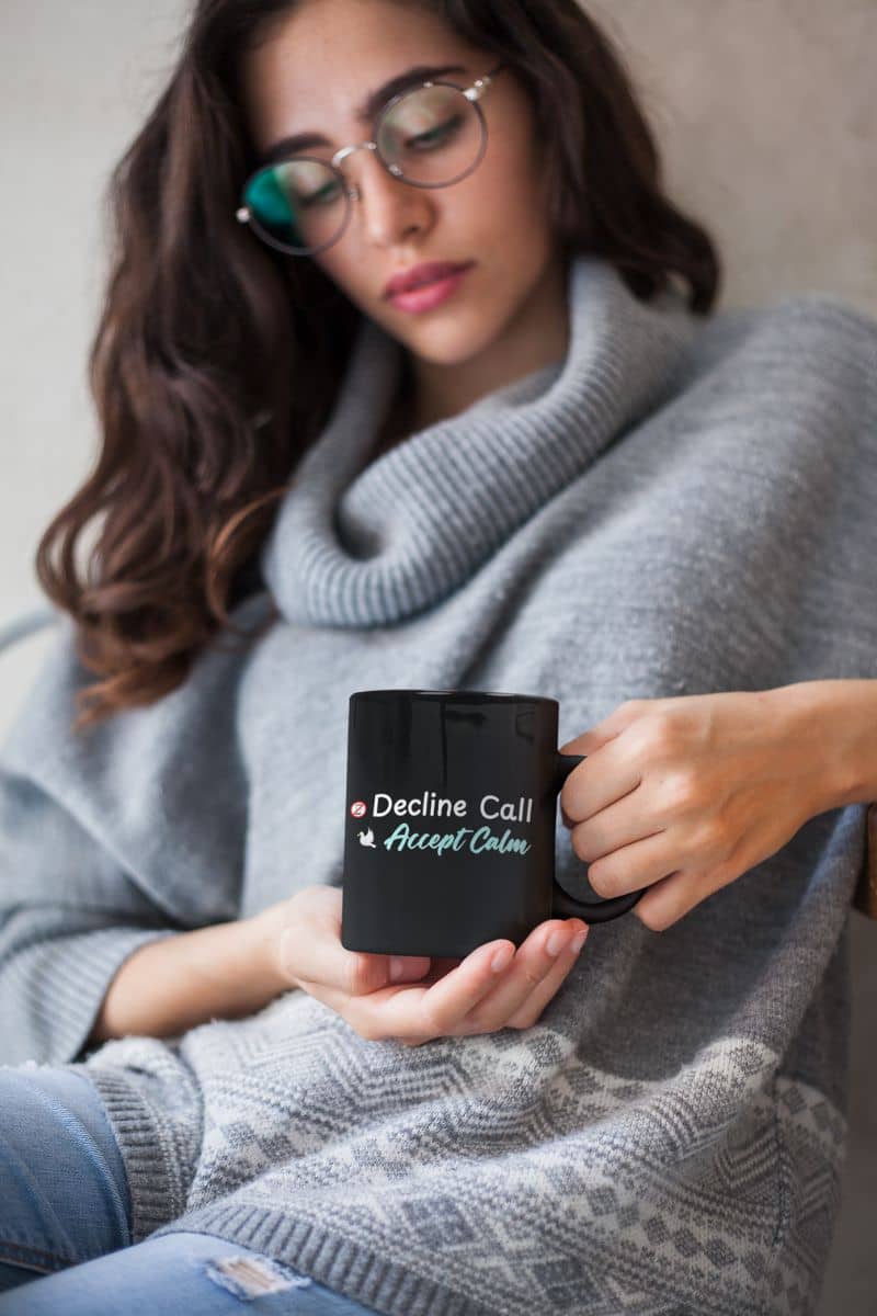 Zen-inspired coffee cup with a woman holding a black mug with text 'Decline Call Accept Calm', wearing a gray sweater and glasses.