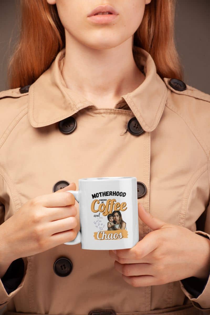 busy mom coffee mug with a person holding a mug with 'Motherhood Powered by Coffee and Chaos' text, wearing a beige coat.