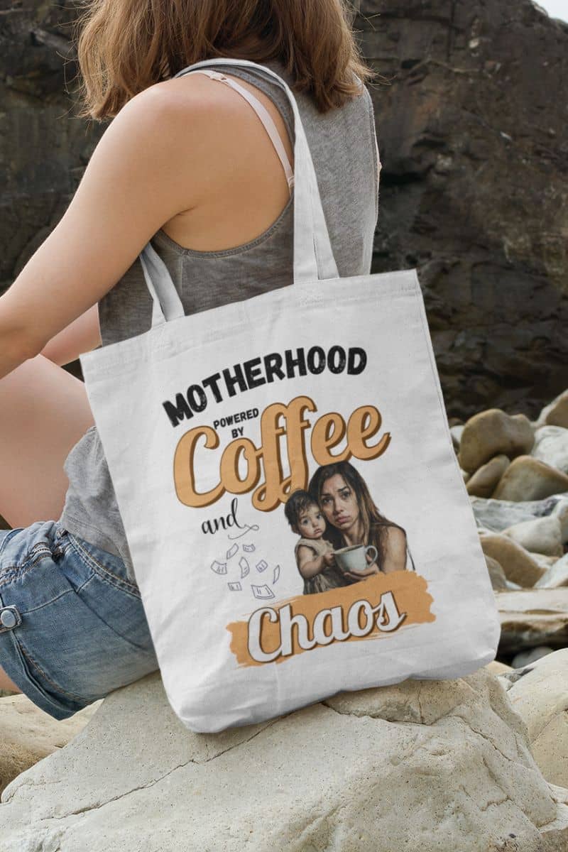 busy mom tote with a person holding a tote bag with 'Motherhood powered by Coffee and Chaos' text and graphic on a rocky surface.