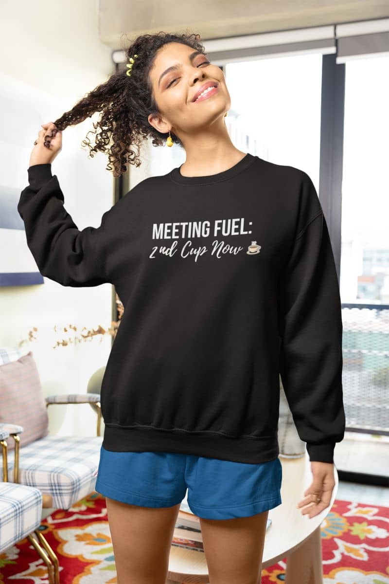 caffeine crewneck with a woman wearing a black sweatshirt with text 'Meeting Fuel: 2nd Cup Now' in a living room