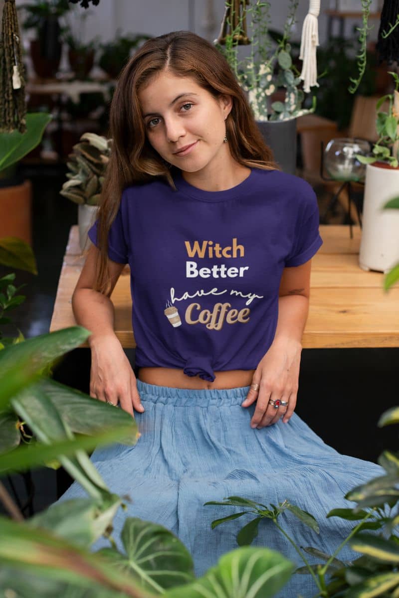 caffeine lover tee with a woman wearing a purple t-shirt with text 'Witch Better Have My Coffee', sitting among plants