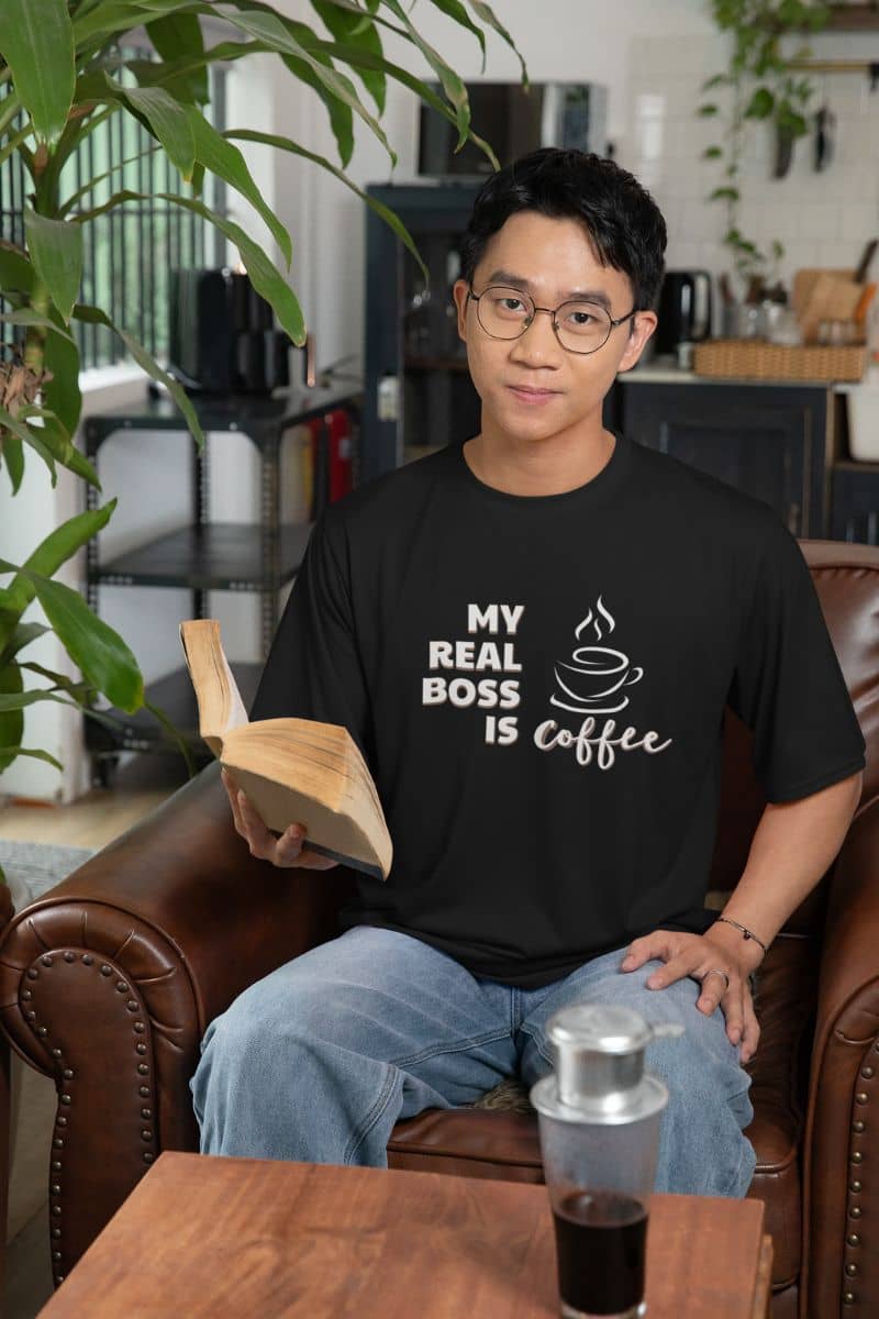 coffee quote t-shirt with a person wearing a black t-shirt with 'My Real Boss is Coffee' text, holding a book, sitting on a brown leather chair.