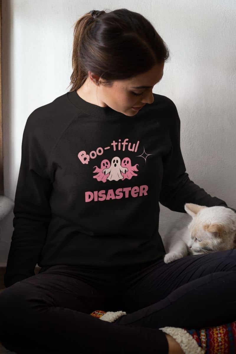 cozy halloween sweatshirt with a person wearing a black sweatshirt with 'Boo-tiful Disaster' text and a cat sitting next to them.