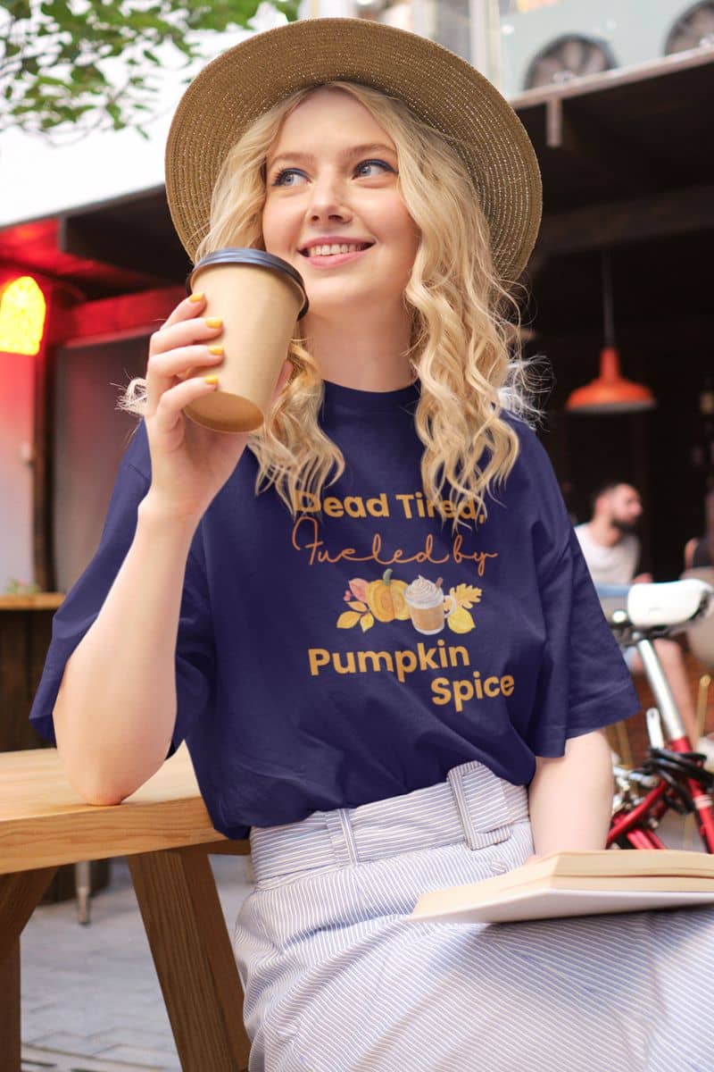 fall coffee shirt with a girl in a blue shirt with text 'Dead Tired, Fueled by Pumpkin Spice', holding a coffee cup outdoors.