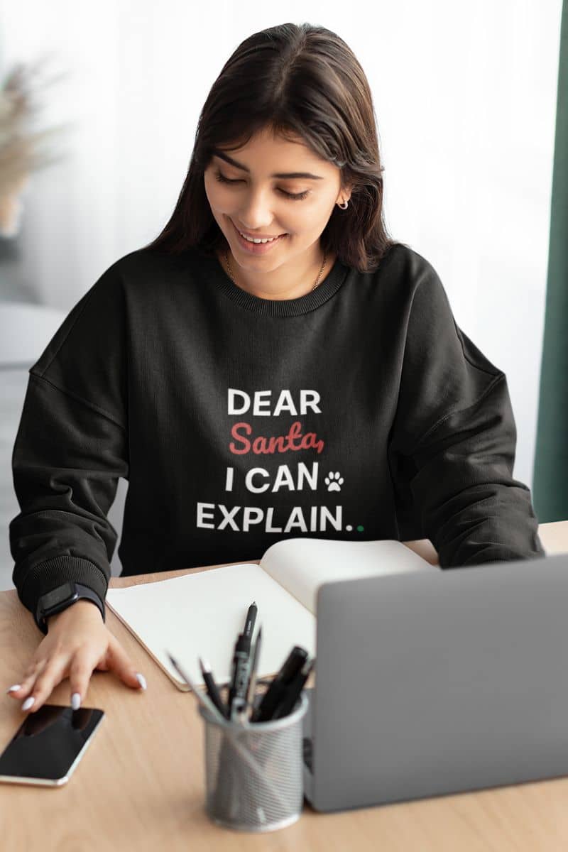 festive sweatshirt with a beautiful girl wearing a black sweatshirt with text 'Dear Santa, I Can Explain', sitting at a desk with a laptop and phone.
