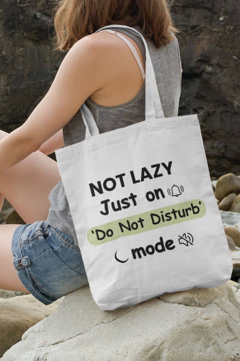 funny introvert bag with a person holding a tote bag with humorous text 'NOT LAZY, Just on 'Do Not Disturb' mode' on a rocky surface