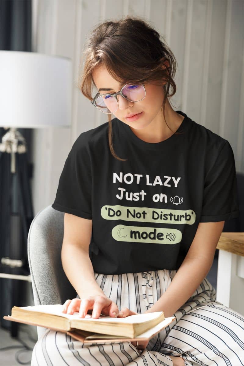funny mental health tee with a woman wearing a black t-shirt with text 'NOT LAZY, Just on 'Do Not Disturb' mode', reading a book indoors.