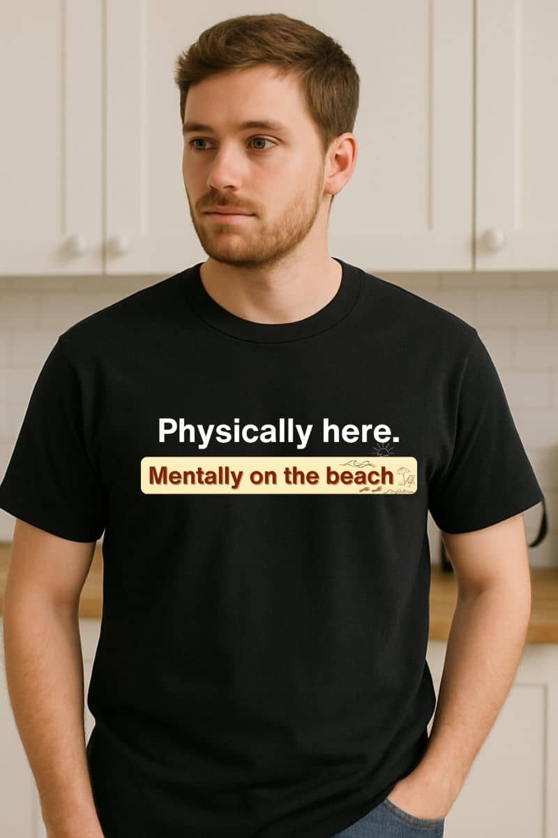 funny summer vacation shirt with a man wearing a black t-shirt with humorous text 'Physically here. Mentally on the beach' in a kitchen setting
