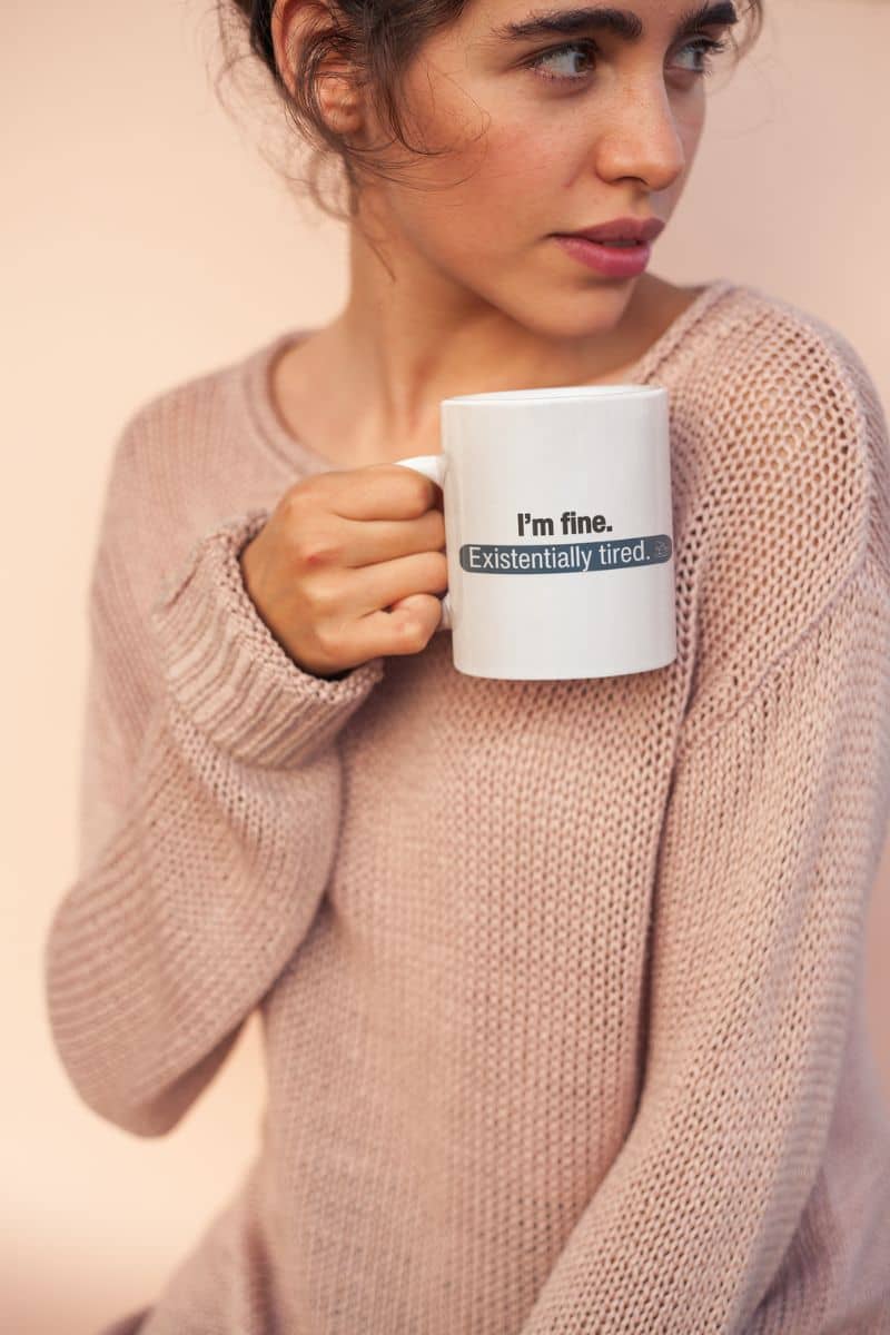 gift for mom coffee mug with a woman holding a mug with text 'I’m fine. Just existentially tired' on it against a plain background