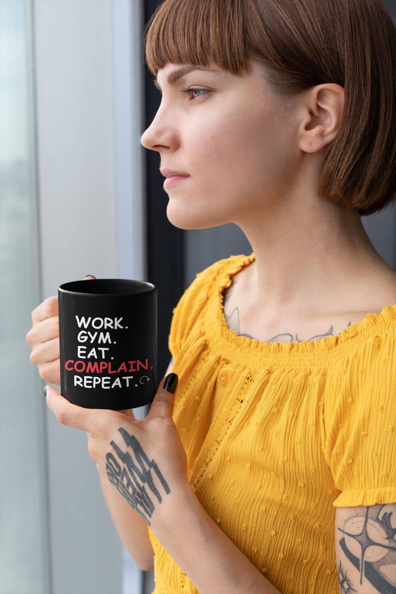 humor ceramic mug with a woman holding a black mug with text 'Work. Gym. Eat. Complain. Repeat', wearing a yellow top.