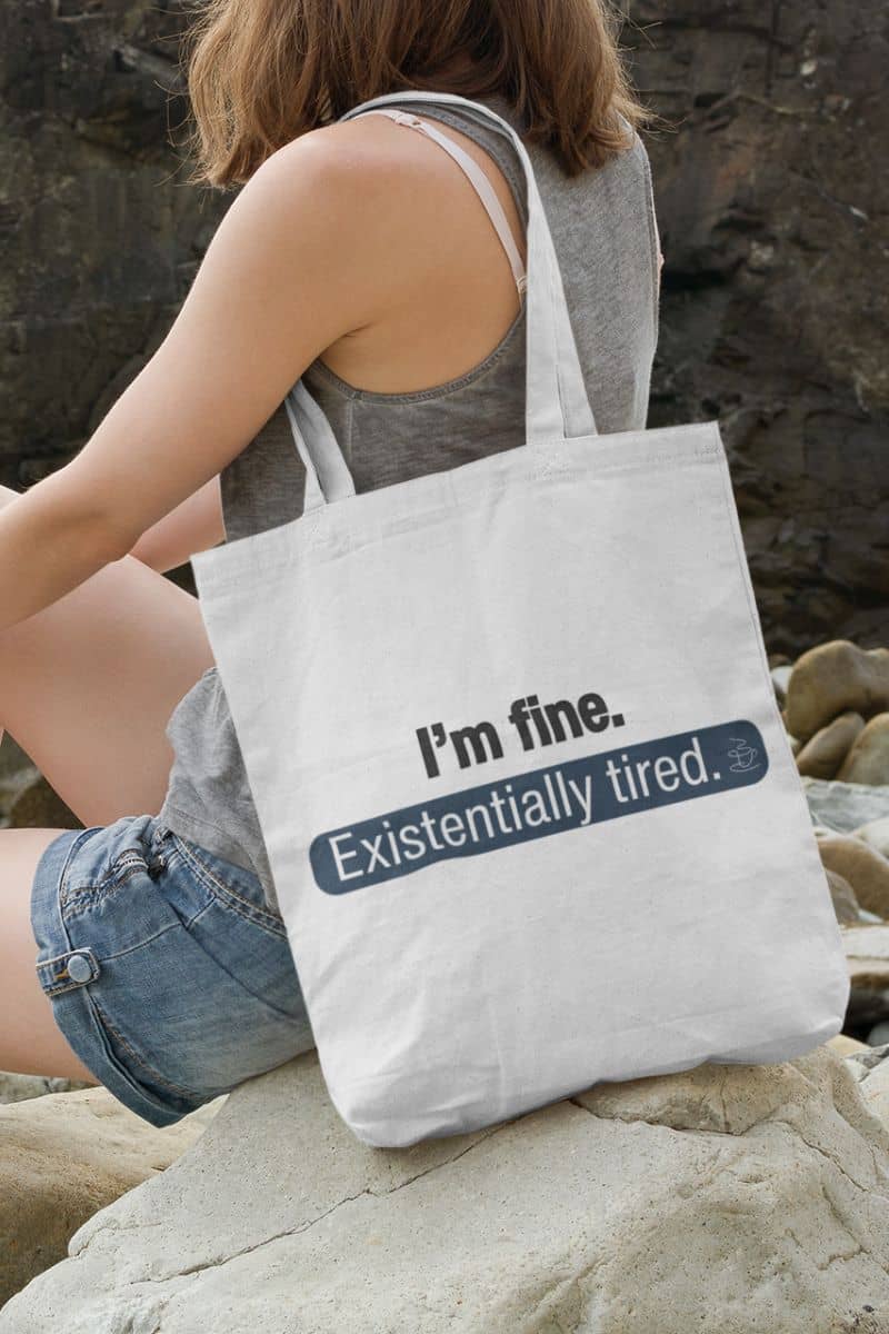 introvert carryall bag with a person holding a tote bag with humorous text 'I’m fine. Just existentially tired' on a rocky surface