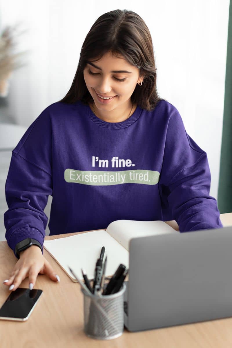 introvert cozy top with a person wearing a purple sweatshirt with text 'I’m fine. Just existentially tired', sitting at a desk with a laptop and phone.