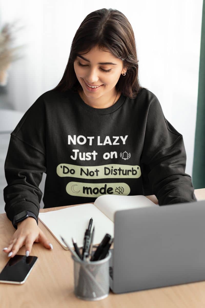 introvert mode sweatshirt with a woman wearing a black sweatshirt with humorous text 'NOT LAZY, Just on 'Do Not Disturb' mode' at a desk with a laptop and phone.