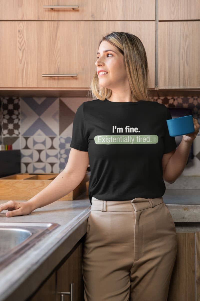 minimalist text tee with a woman in a kitchen wearing a black t-shirt with text 'I’m fine. Just existentially tired', holding a blue mug.