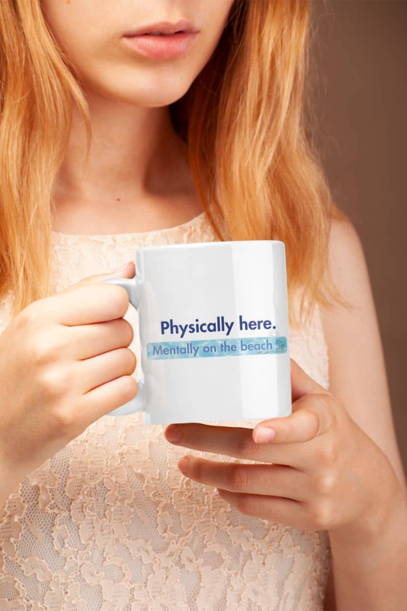 ocean theme ceramic mug with a person holding a mug with text 'Physically here, mentally on the beach' against a neutral background