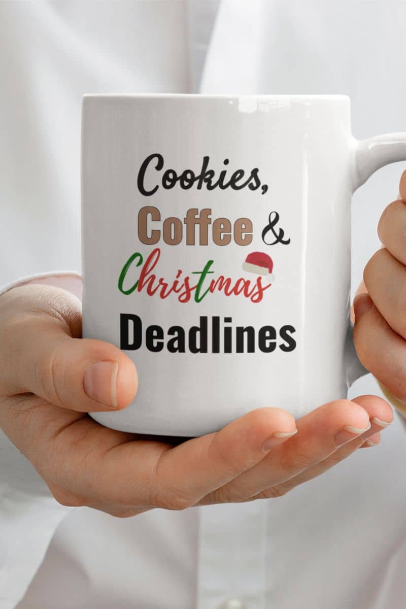 office coffee mug with a white mug with 'Cookies, Coffee & Christmas Deadlines' text held by a person.