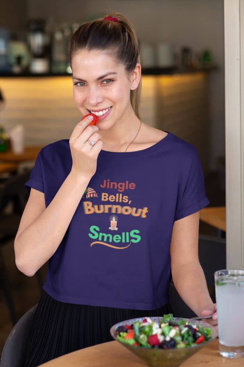 office humor t-shirt with a woman wearing a purple t-shirt with humorous text 'Jingle Bells, Burnout Smells', eating a red lollipop in a casual setting.