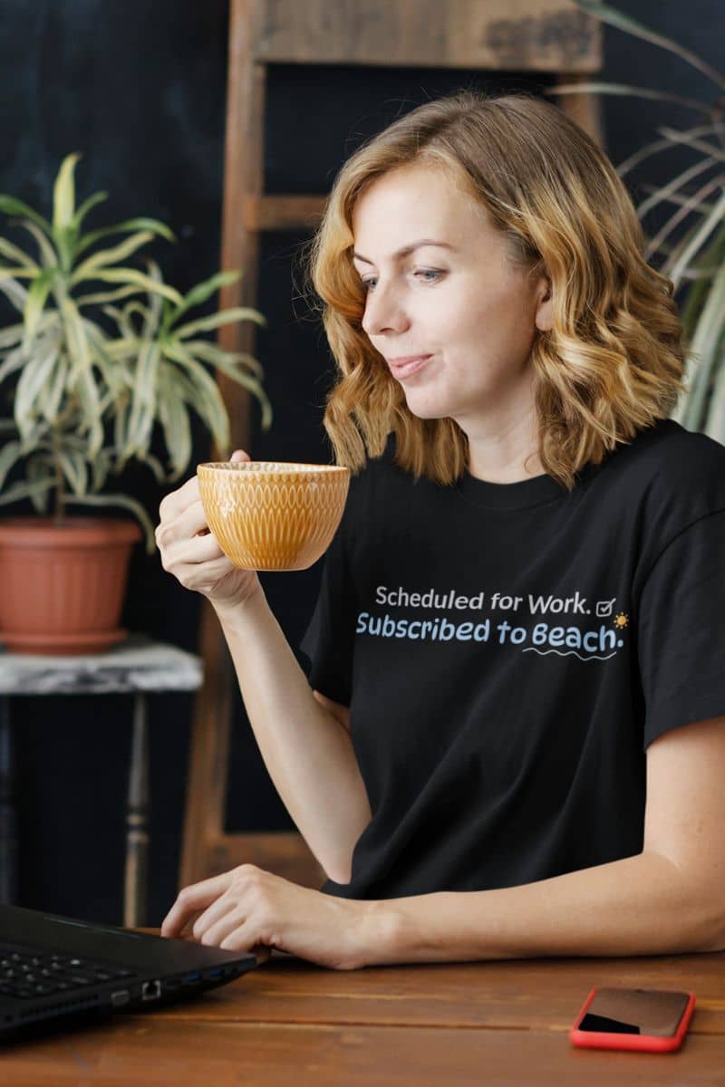 oversized beach t-shirt with a woman holding a cup with text on her shirt, sitting at a desk with a laptop and phone.