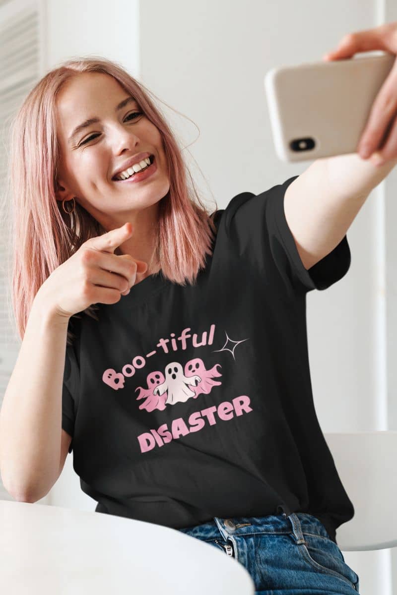 pumpkin graphic tee with a girl wearing a black t-shirt with text 'Boo-tiful Disaster' and graphics, taking a selfie.