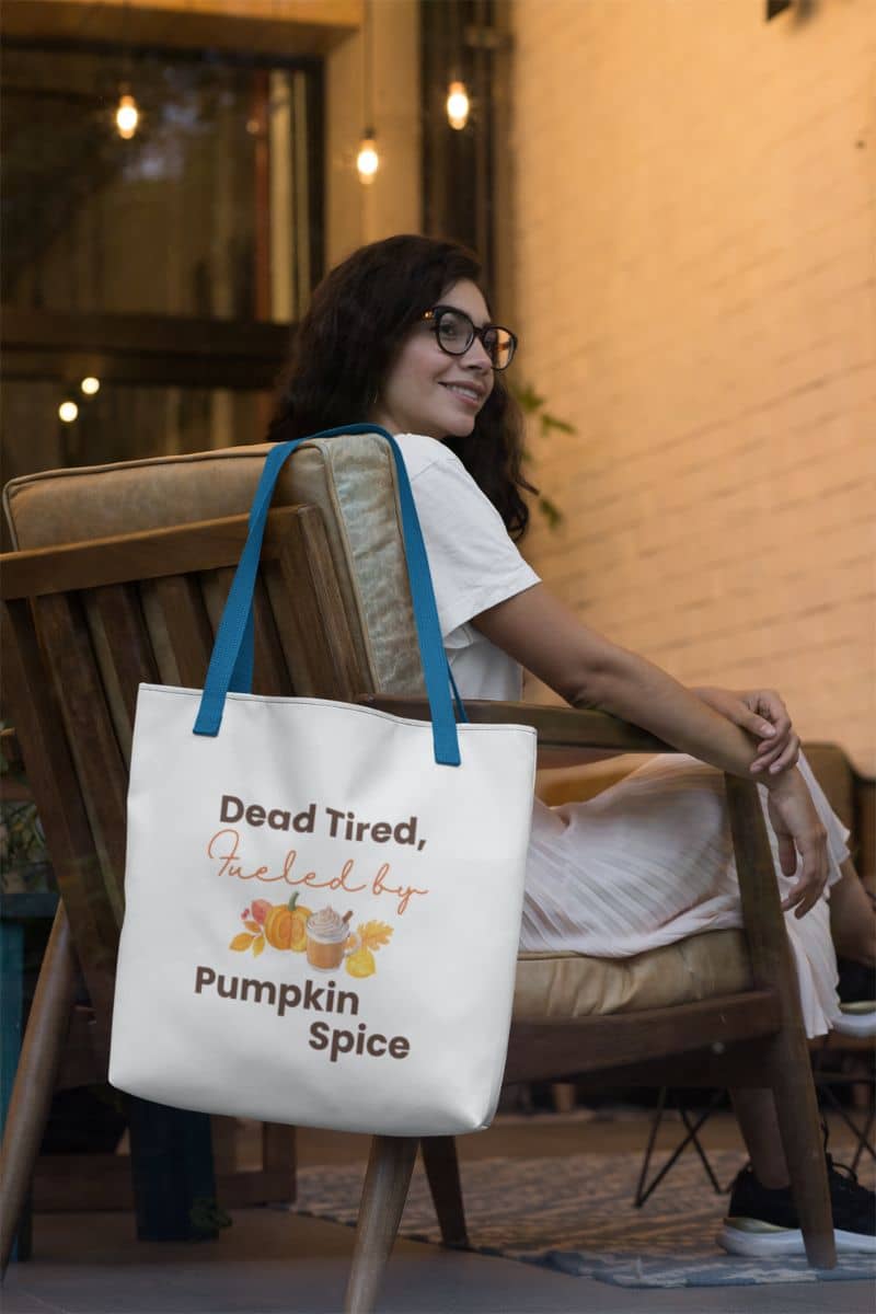 pumpkin spice bag with a woman sitting on a chair with a tote bag featuring 'Dead Tired, Fueled by Pumpkin Spice' text.