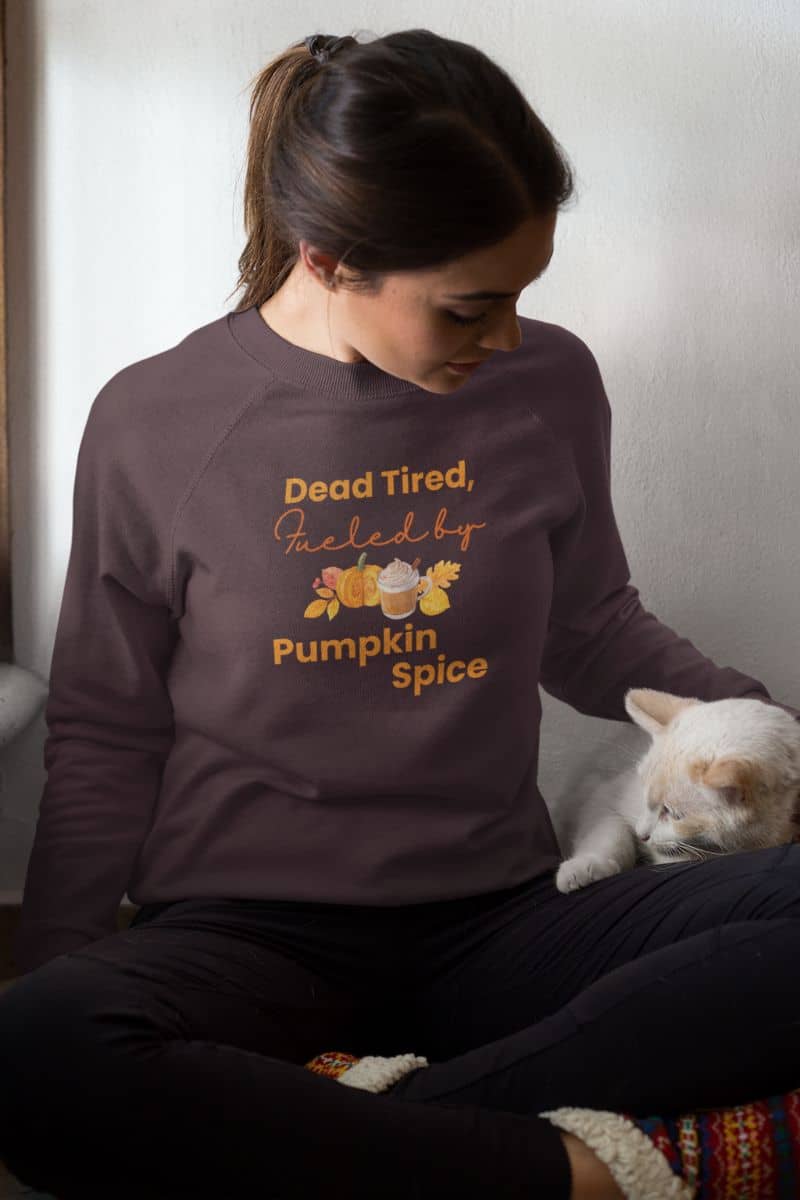 pumpkin spice sweatshirt with a person wearing a sweatshirt with 'Dead Tired, Fueled by Pumpkin Spice' text sitting with a cat.