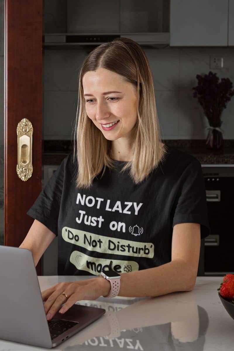 quiet life tee with a woman wearing a black t-shirt with text 'NOT LAZY, Just on 'Do Not Disturb' mode', using a laptop in a kitchen.