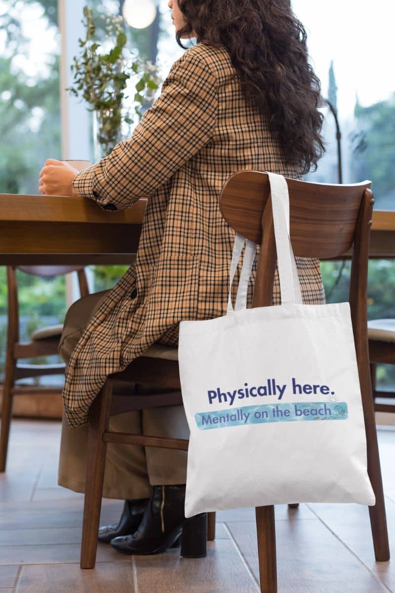 sarcastic travel tote with a person sitting at a table with a tote bag that says 'Physically here. Mentally on the beach.'