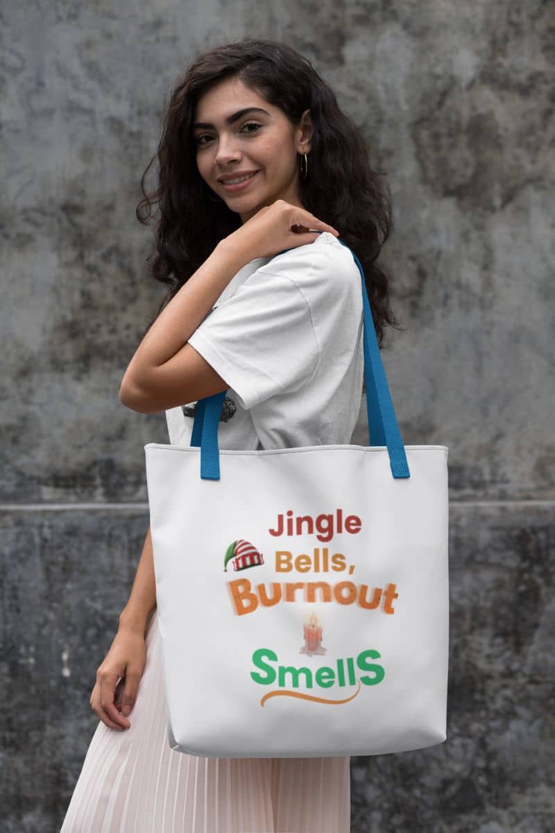 sarcastic work tote with a woman holding a tote bag with humorous text 'Jingle Bells, Burnout Smells' against a gray wall.