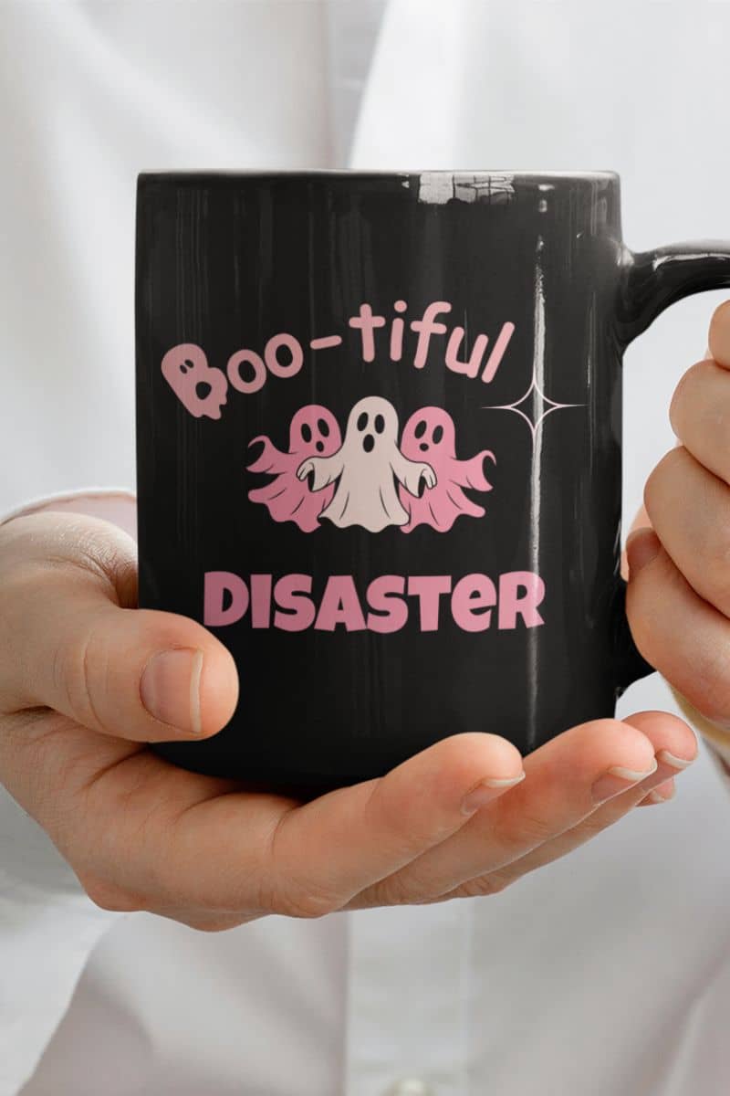 scary but cute coffee mug with a black mug with 'Boo-tiful Disaster' text and ghost graphics held by hands against a white background