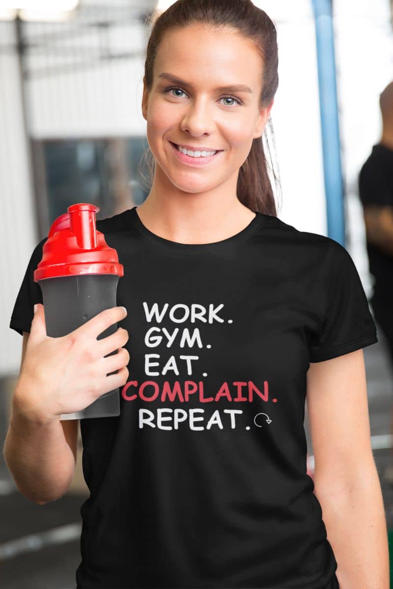 unisex graphic shirt with a woman wearing a black t-shirt with text 'Work. Gym. Eat. Complain. Repeat', holding a shaker bottle in a gym setting.