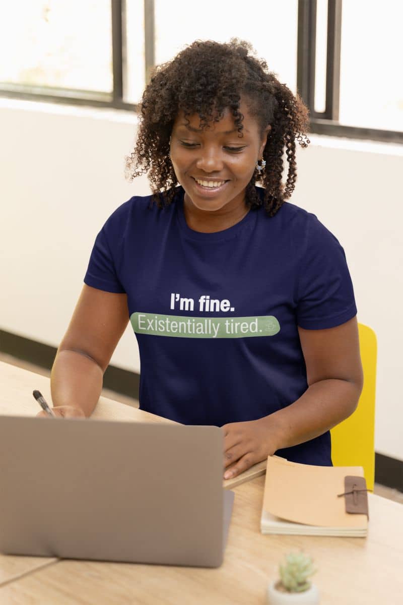 witty existential shirt with a person wearing a blue t-shirt with humorous text 'I’m fine. Just existentially tired', sitting at a desk with a laptop.