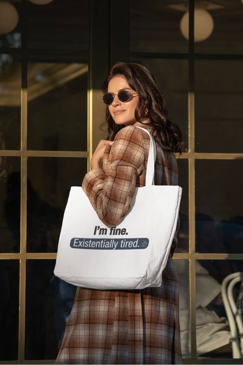 witty quote tote with a woman holding a tote bag with text 'I’m fine. Just existentially tired' in front of a glass window