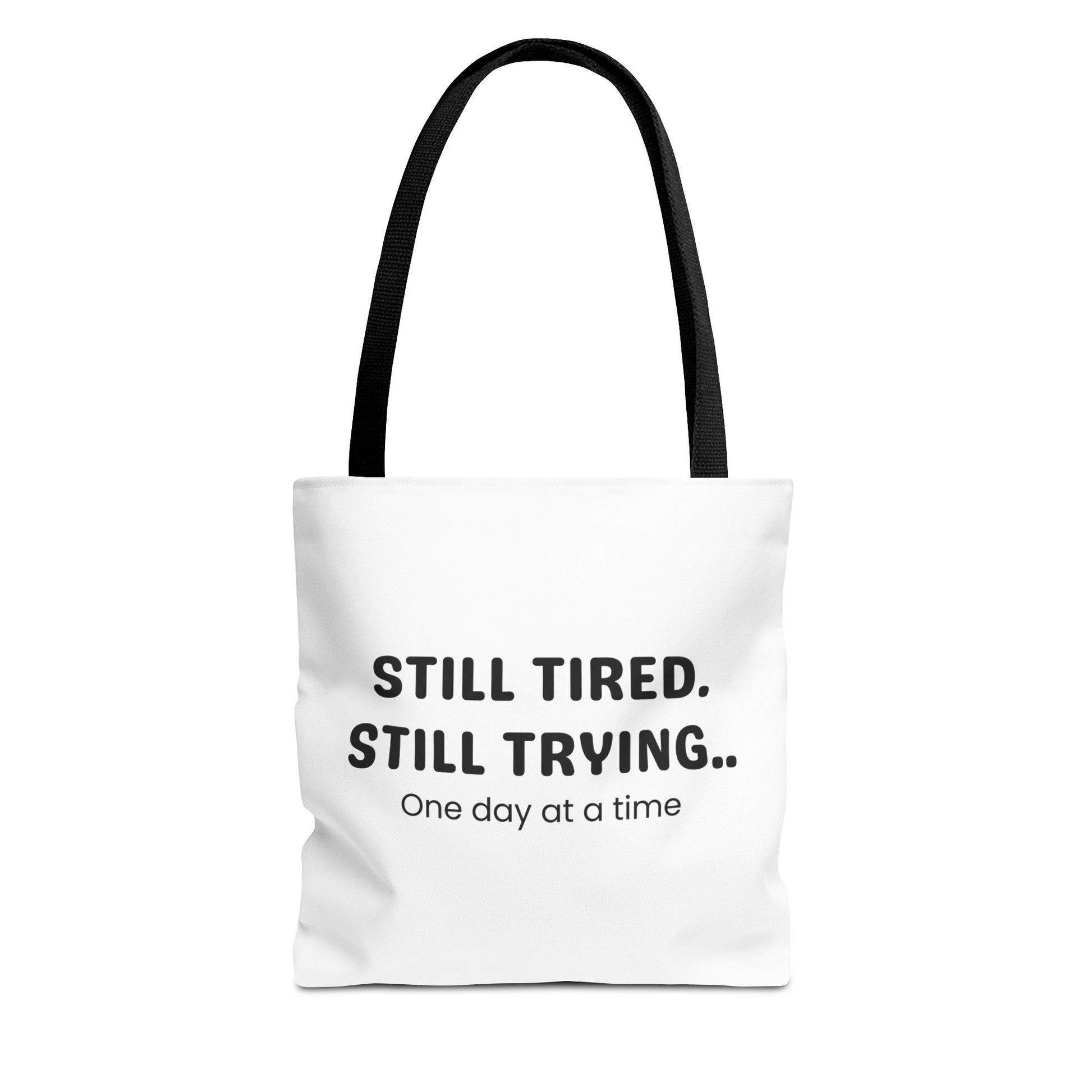 Tote bag with black handle and 'Still Tired. Still Trying.. One day at a time' text on a white background