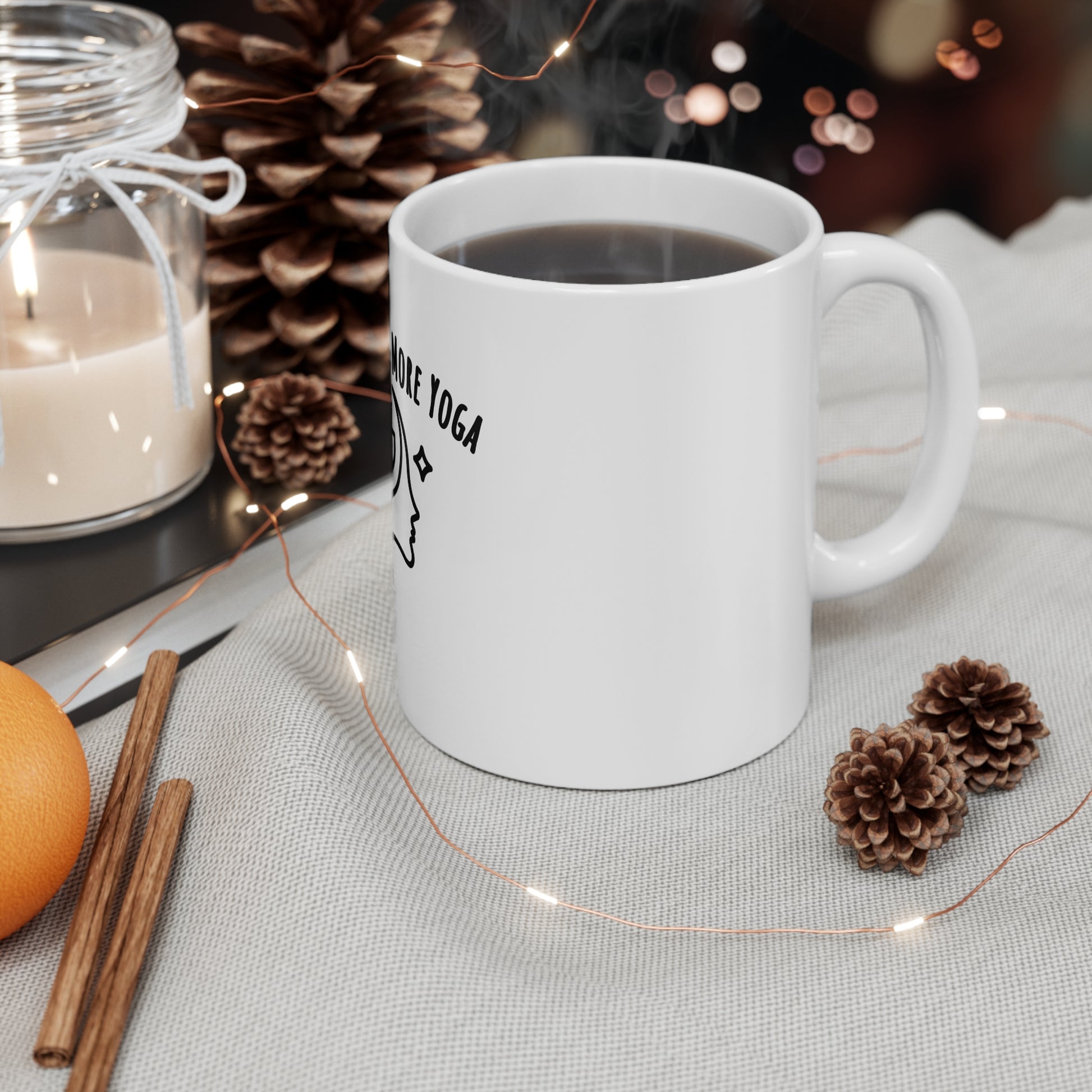 White mug with text 'Less Talk, More Yoga' on a table with a candle, orange, and pine cones.