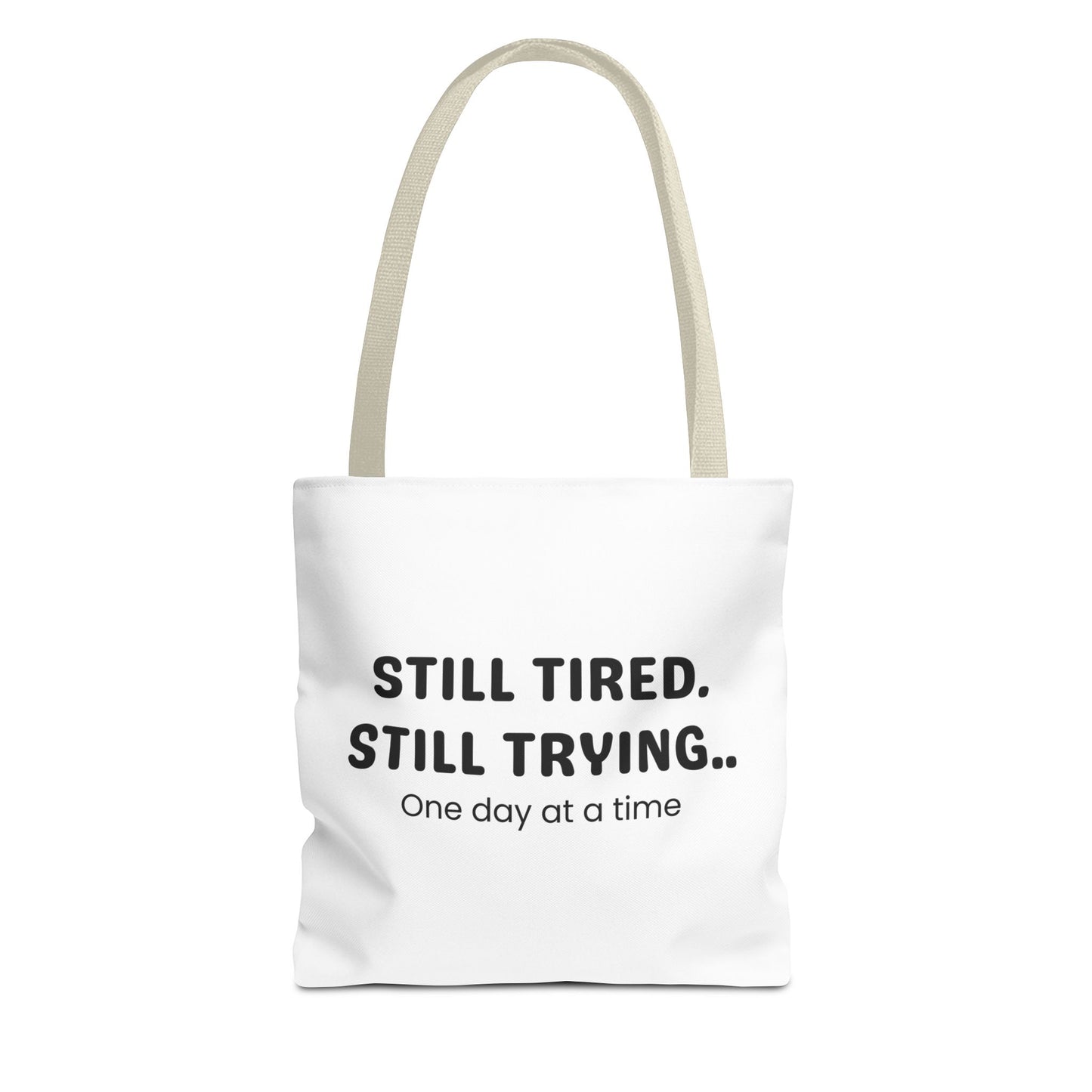Tote bag with beige handle and 'Still Tired. Still Trying.. One day at a time' text on a white background