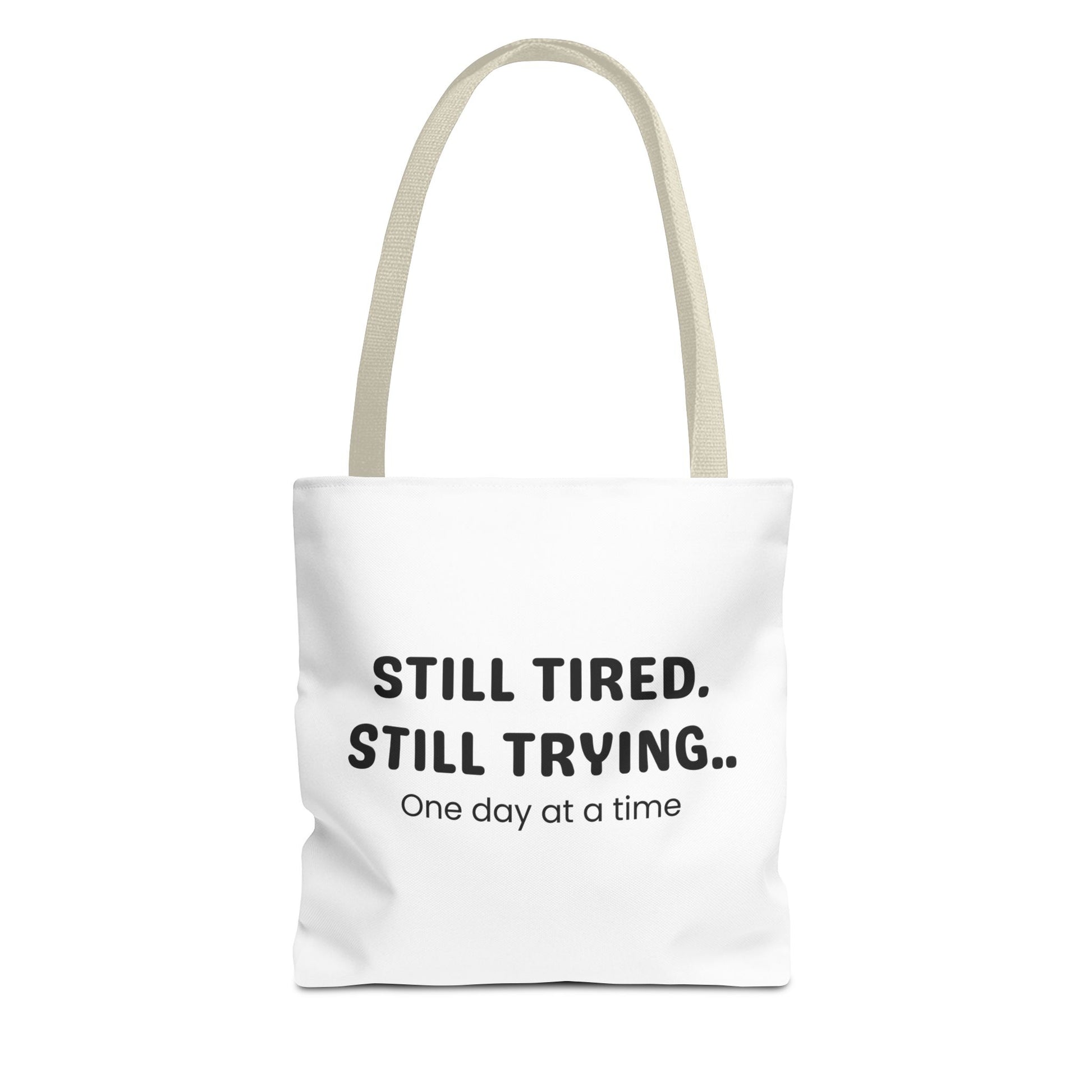 Tote bag with beige handle and 'Still Tired. Still Trying.. One day at a time' text on a white background
