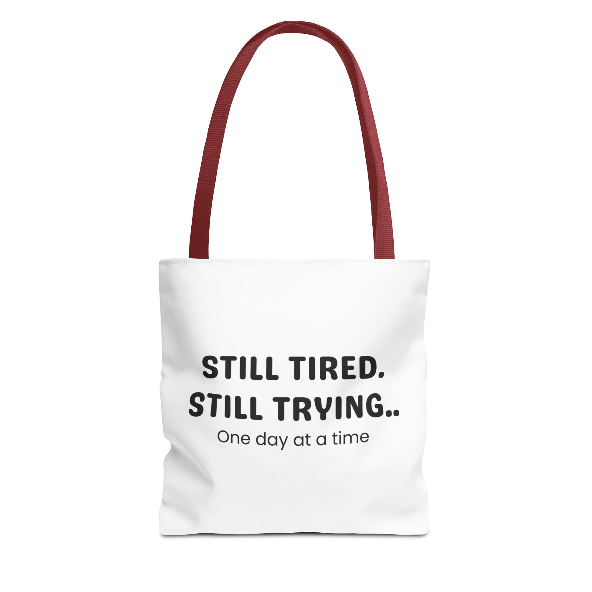 Tote bag with red handle and 'Still Tired. Still Trying.. One day at a time' text on a white background