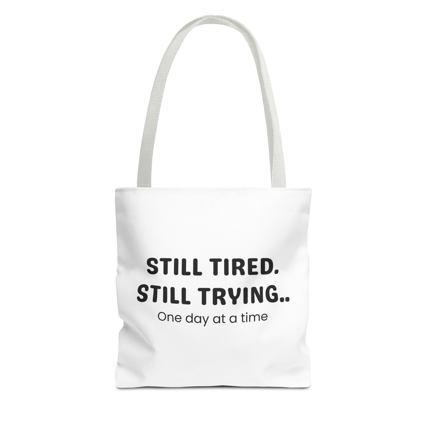 Tote bag with white handle and 'Still Tired. Still Trying.. One day at a time' text on a white background