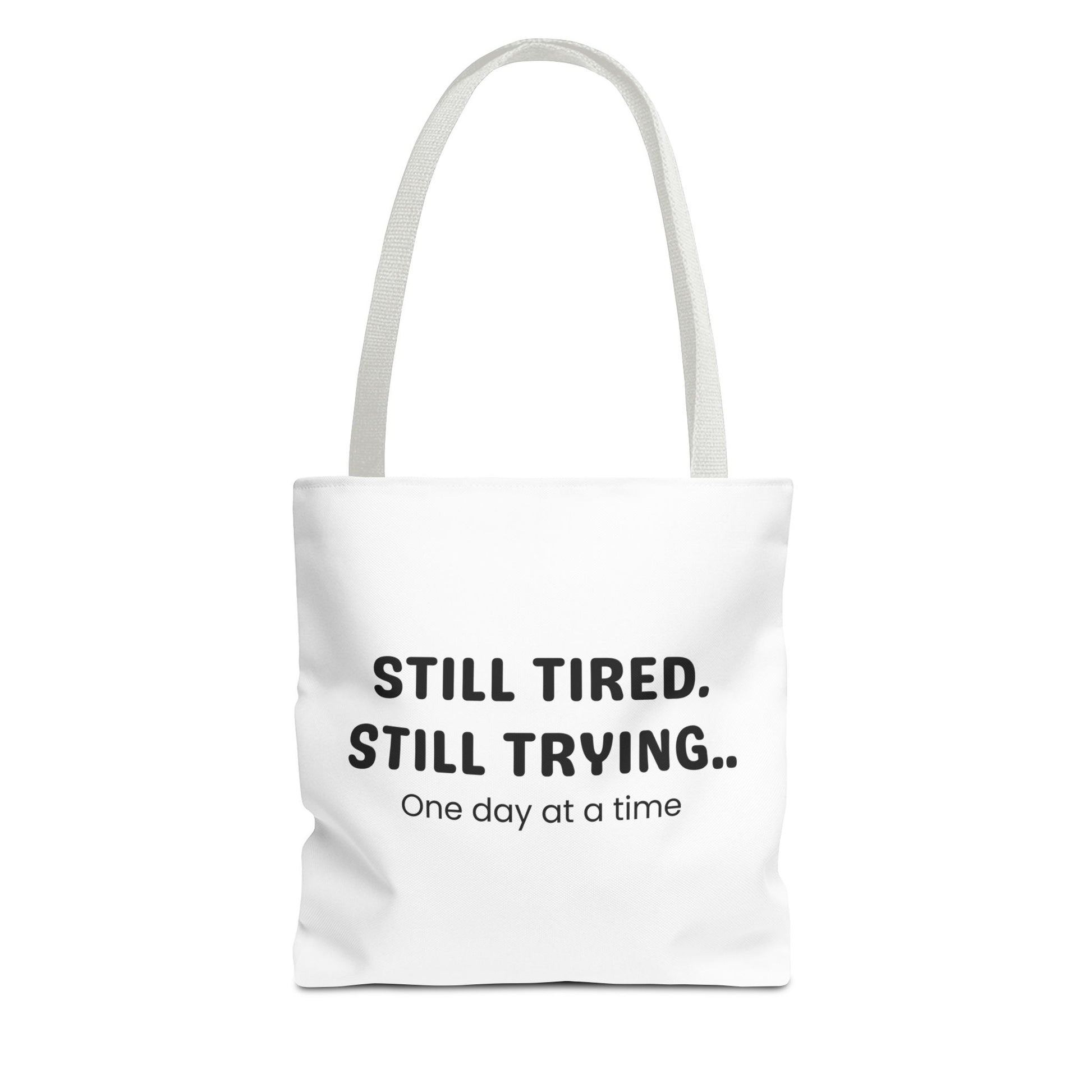 Tote bag with white handle and 'Still Tired. Still Trying.. One day at a time' text on a white background