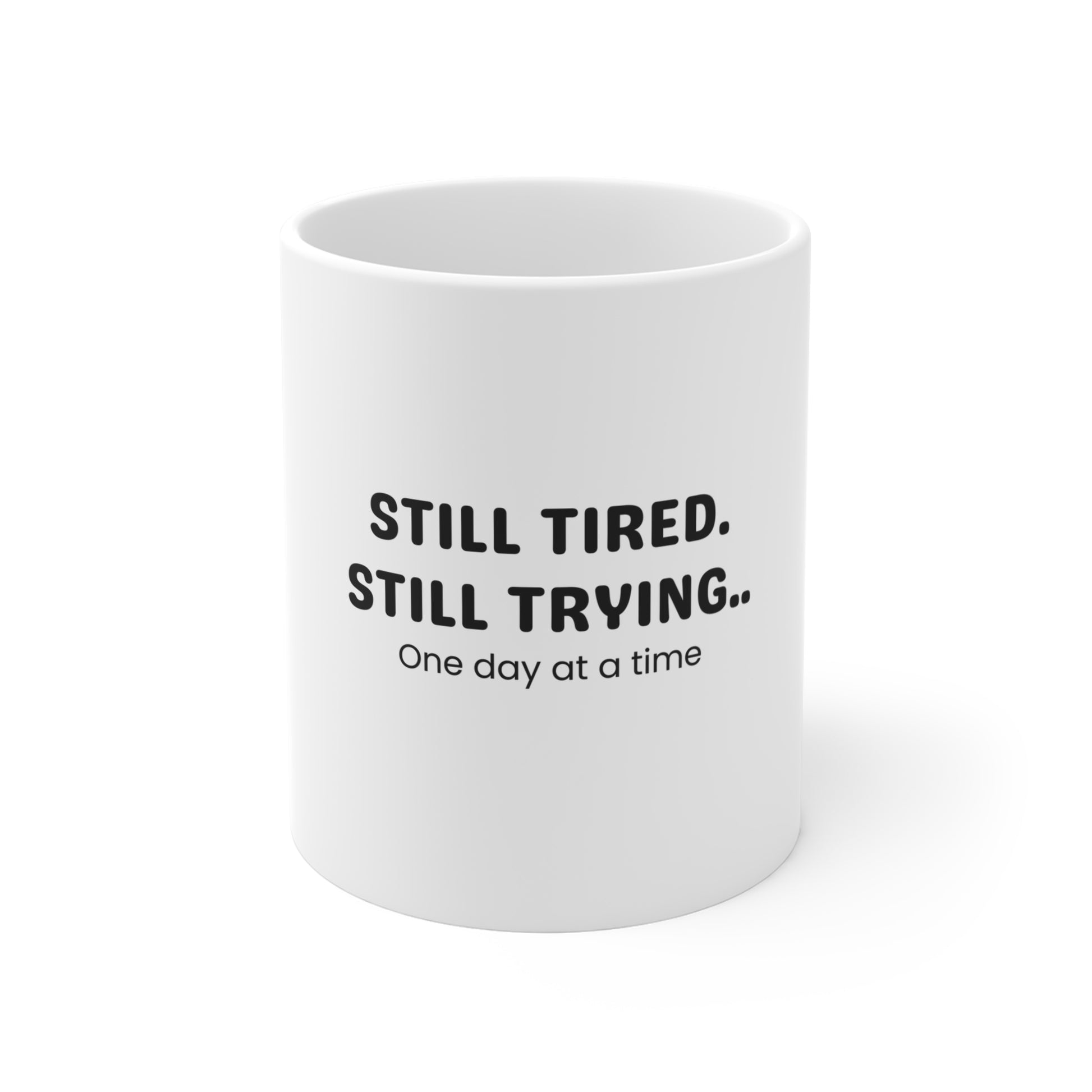 White mug with black text 'STILL TIRED. STILL TRYING.. One day at a time' on a white background