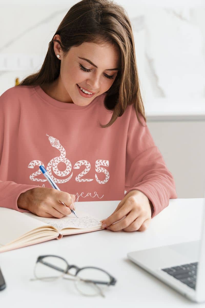 2025: Your Year Sweatshirt with a girl sitting at home working on her laptop