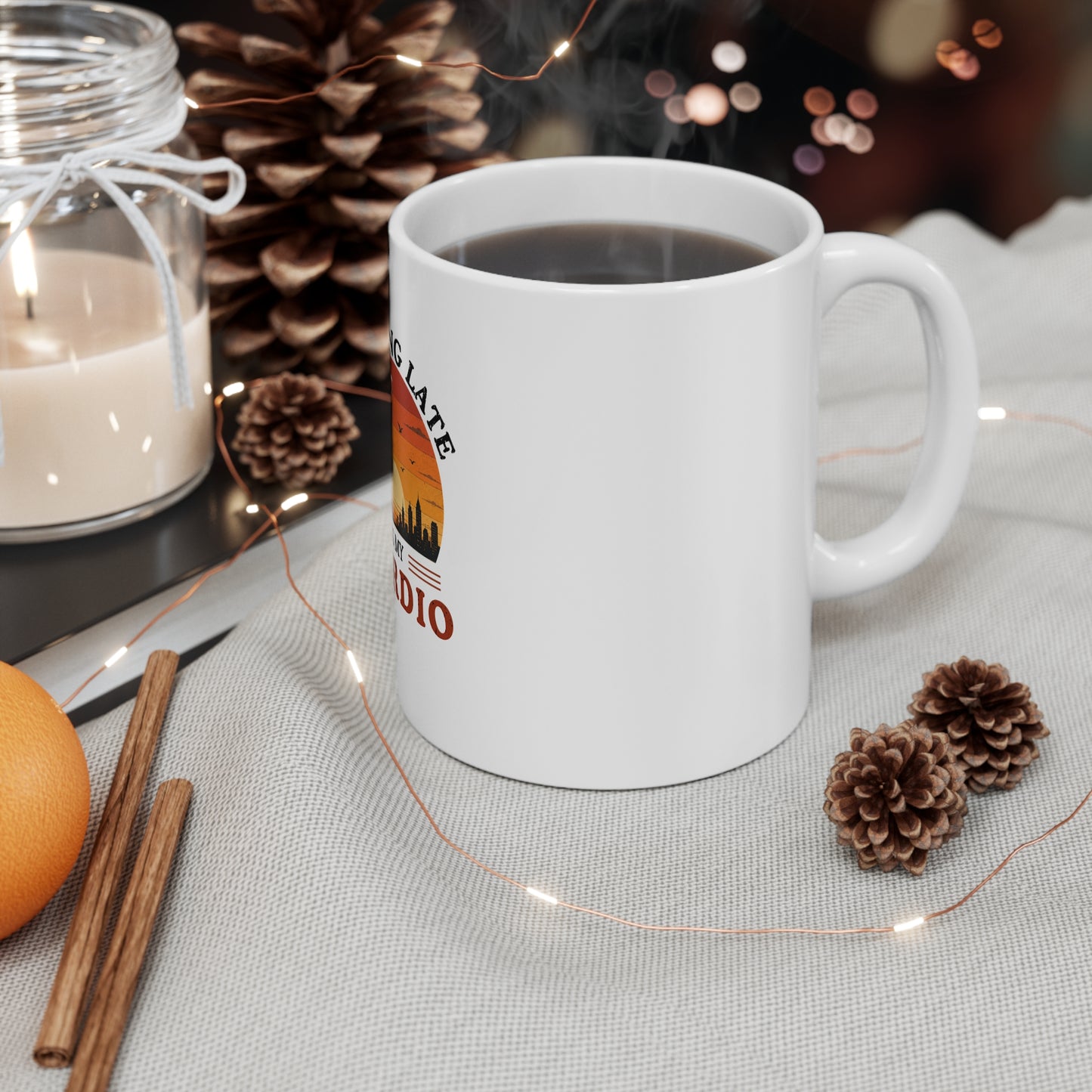 White mug with a coffee design on a table with a candle, orange, and pine cones.