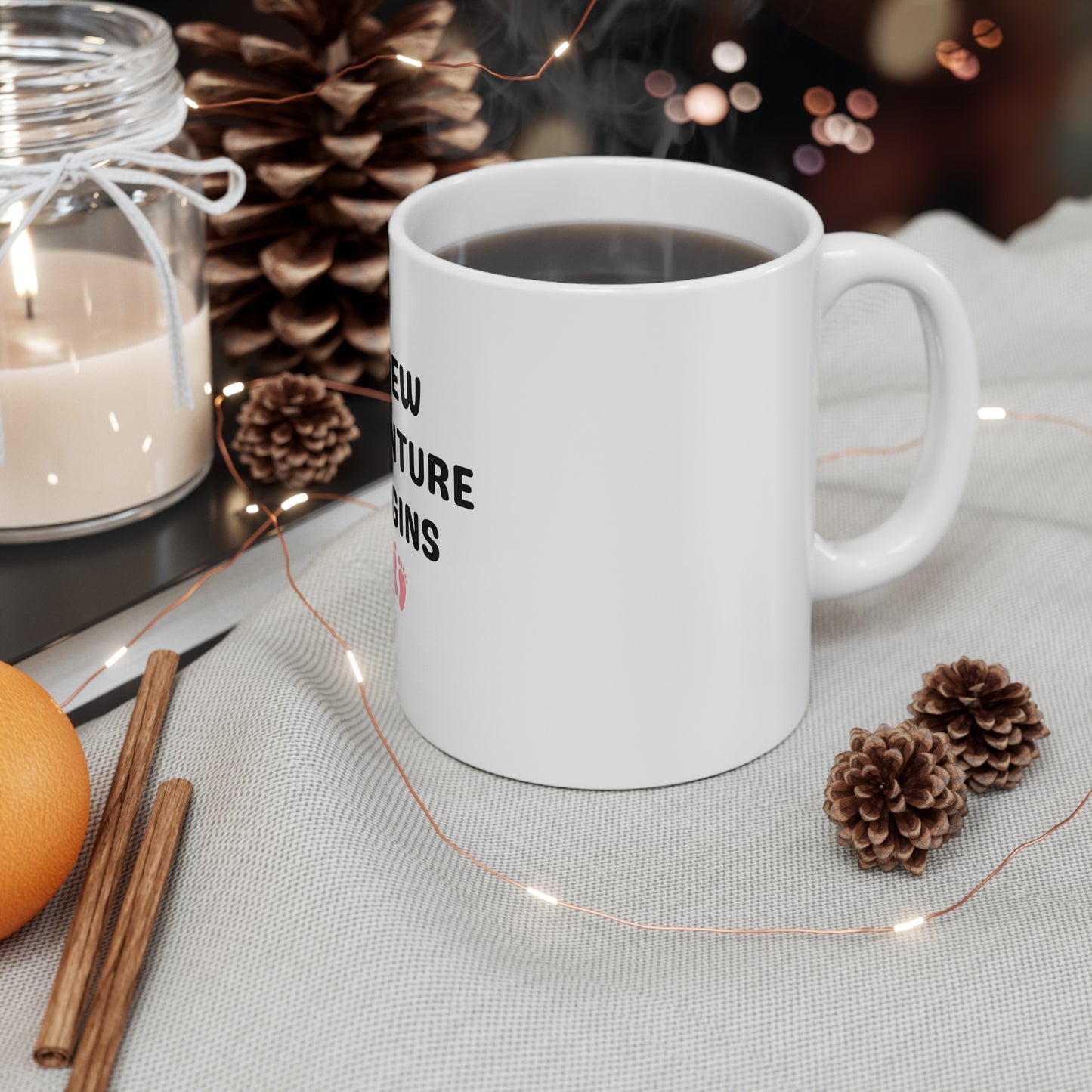White mug with text 'New Adventure Begins' on a table with decorative items including a candle, oranges, and pine cones.