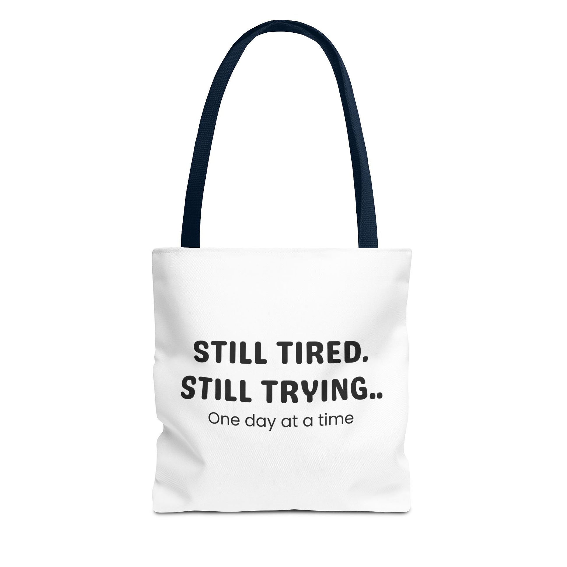 Tote bag with navy handle and 'Still Tired. Still Trying.. One day at a time' text on a white background
