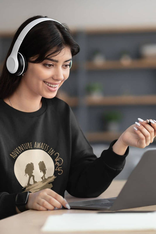 Adventure awaits in 2025 sweatshirt with the girl wearing headphones and looking at the laptop with a bright smile