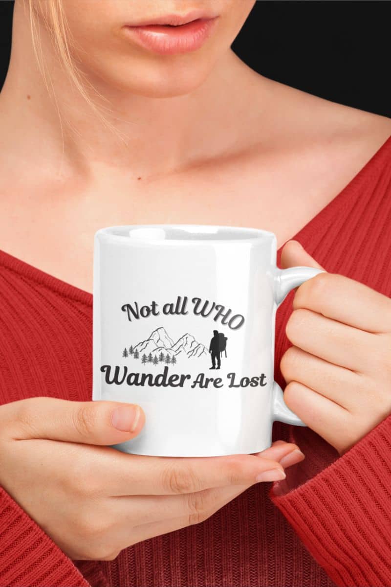 Adventure coffee mug with a person holding a white mug with text 'Not all who wander are lost' and a mountain graphic, wearing a red sweater.