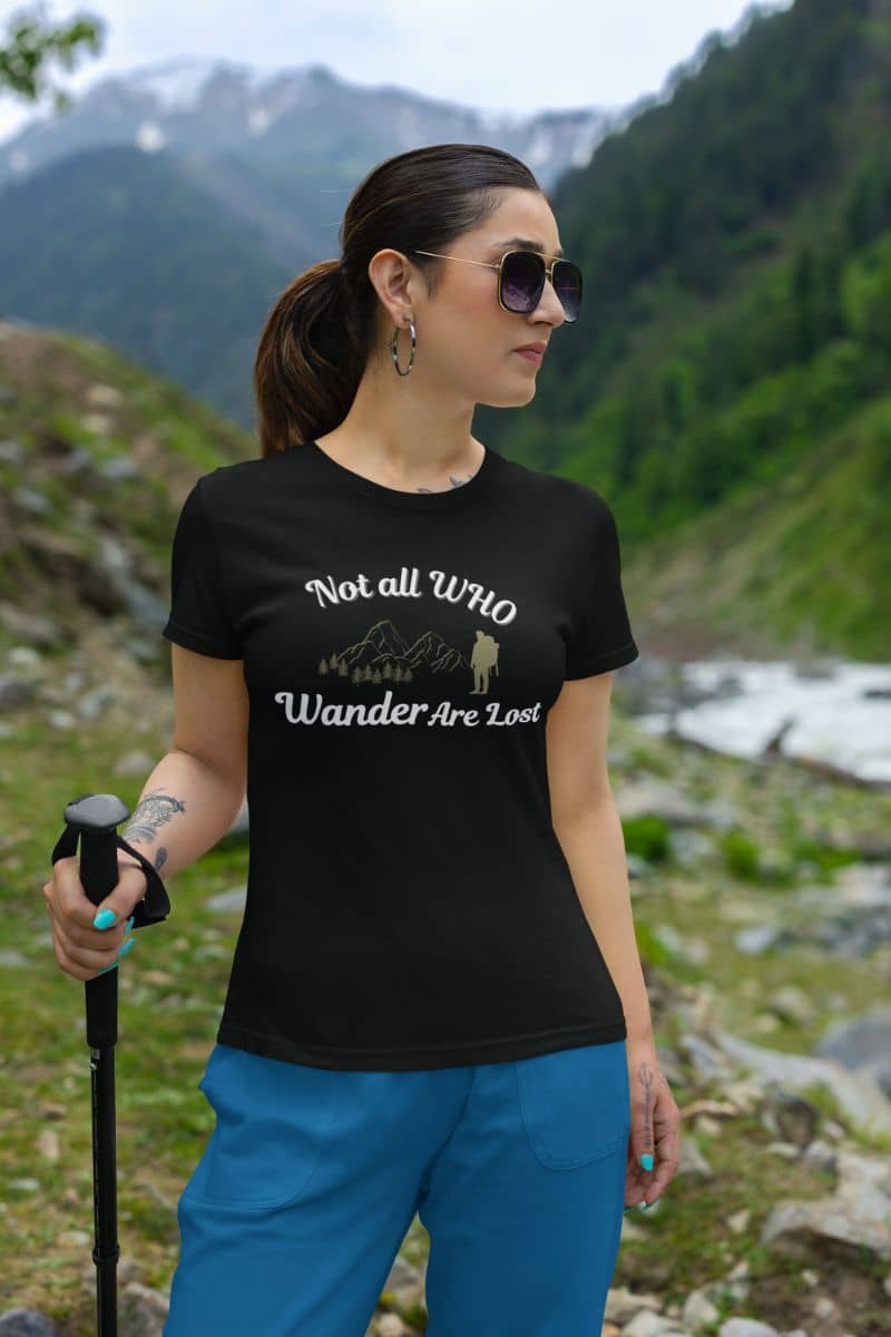 Adventure t-shirt with a woman hiking in a mountainous area wearing a black t-shirt with text 'Not all who wander are lost' and blue pants.