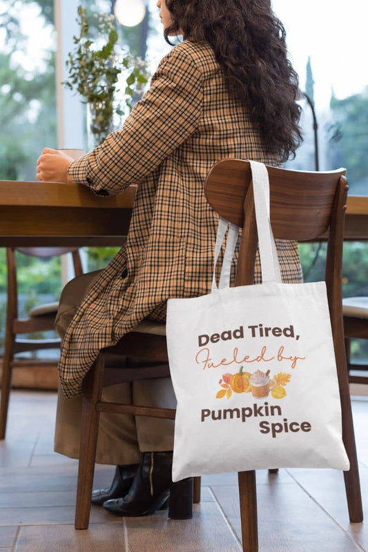 Autumn coffee tote bag with a person sitting at a table with a tote bag that says 'Dead Tired, Fueled by Pumpkin Spice' in a cozy indoor setting.