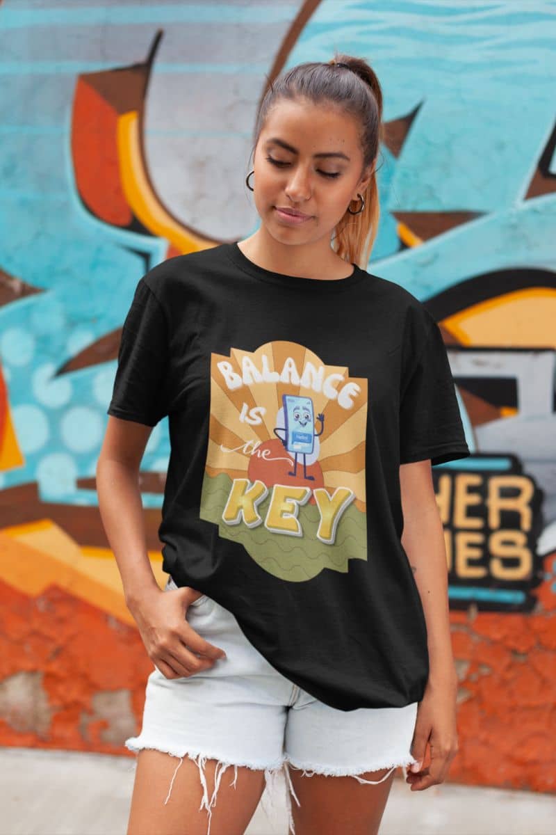 Balance Is The Key T-Shirt with a woman wearing a black t-shirt with a graphic design in front of a colorful mural.