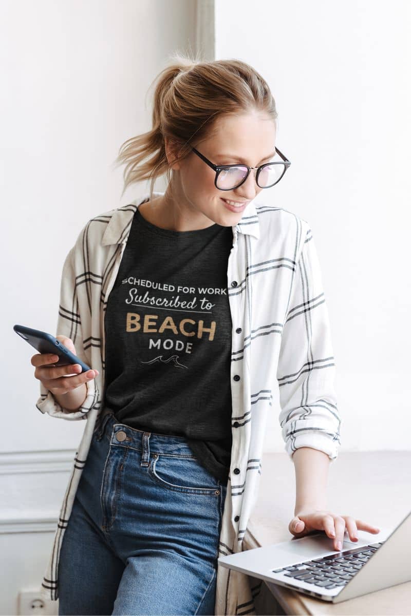 Beach vacation shirt with a woman wearing a black t-shirt with a 'Scheduled for Work. Subscribed to Beach' design, using a laptop.